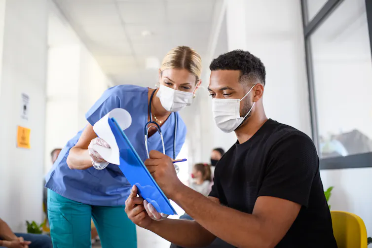 A nurse assists a young man to fill out a form. Both are wearing masks.