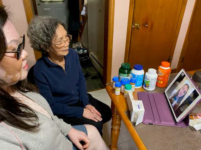 Two women sit at a table, where bottles and boxes of medicines and supplements are crowded near a computer tablet screen, where a pharmacist is meeting with them over videoconferencing.