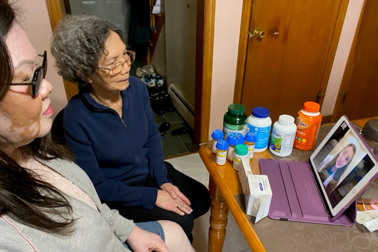 Two women sit at a table, where bottles and boxes of medicines and supplements are crowded near a computer tablet screen, where a pharmacist is meeting with them over videoconferencing.
