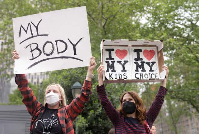Two women stand with signs above their heads, one reads 'my body' the other reads 'i love my kids, i love my choice'