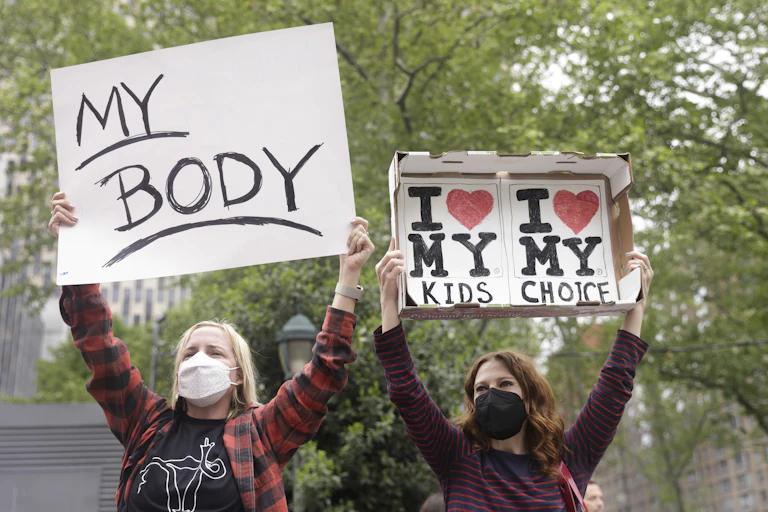 Two women stand with signs above their heads, one reads 'my body' the other reads 'i love my kids, i love my choice'