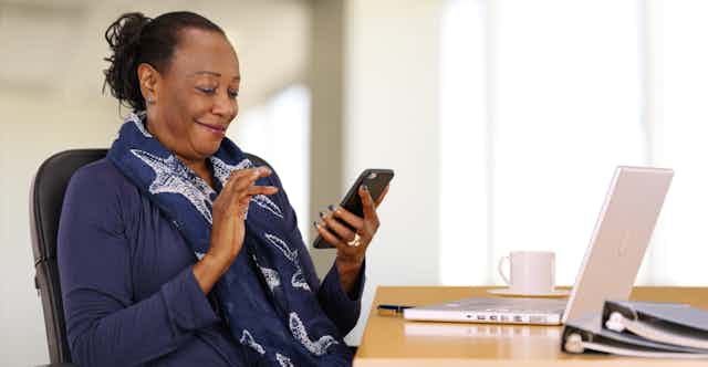 A Black woman sits at a desk looking at her smartphone with a laptop in front of her.