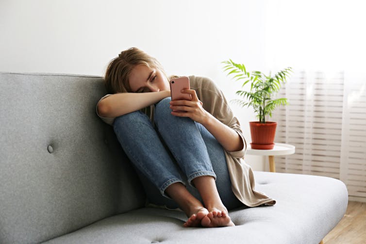 Distressed person curled up on sofa looking at smartphone