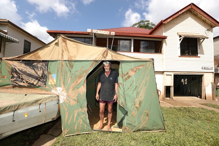 man stands next to tent in front of damaged home