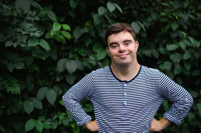 Man with Down syndrome smiling with hands on hips in front of garden bush