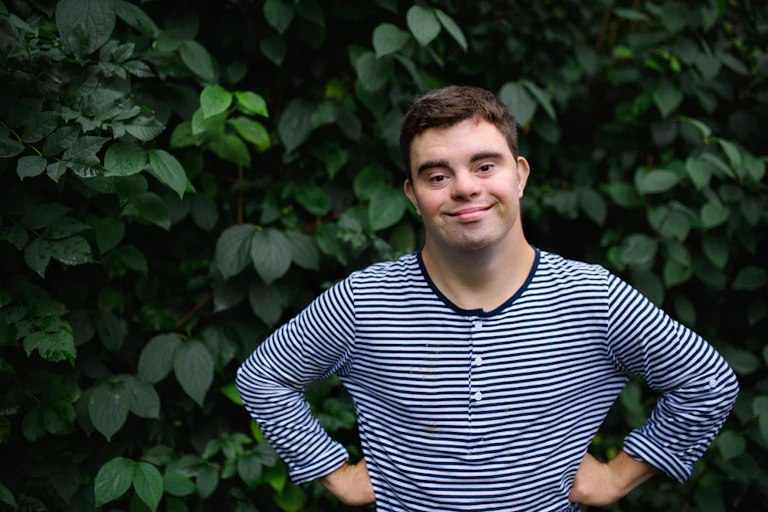 Man with Down syndrome smiling with hands on hips in front of garden bush