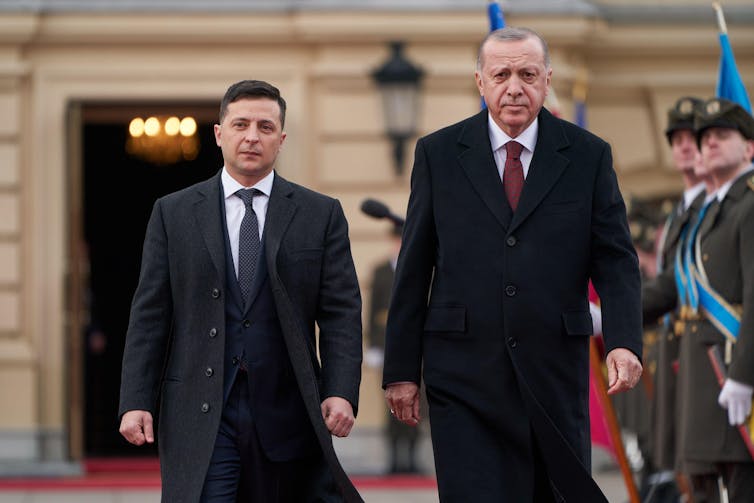 Two men in black coats walk forward on a red carpet.