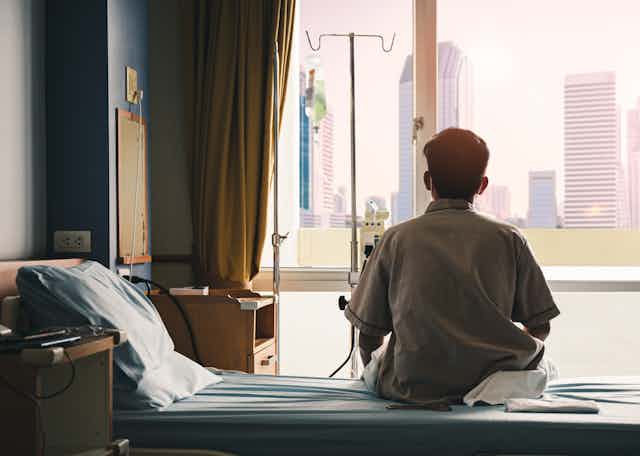 A patient looks out a window while sitting on a bed.