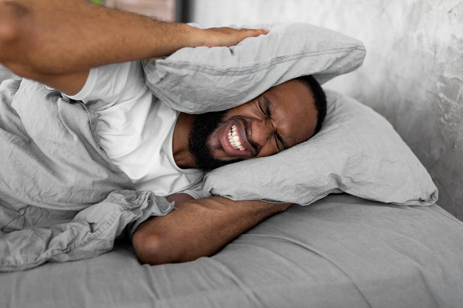 A young man struggles to sleep due to noise. He is holding two pillows over his ears in annoyance.