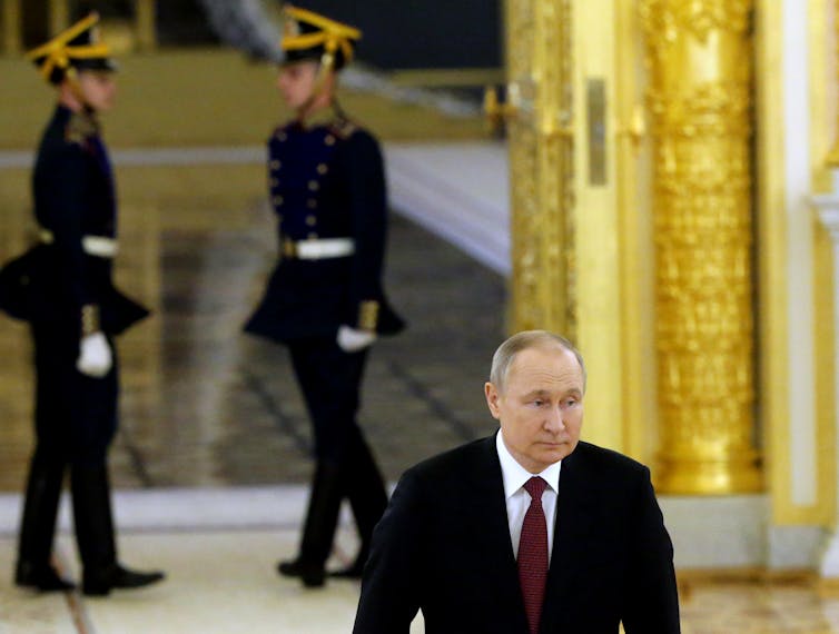 Putin is seen walking through an ornate hall, with two soldiers behind him