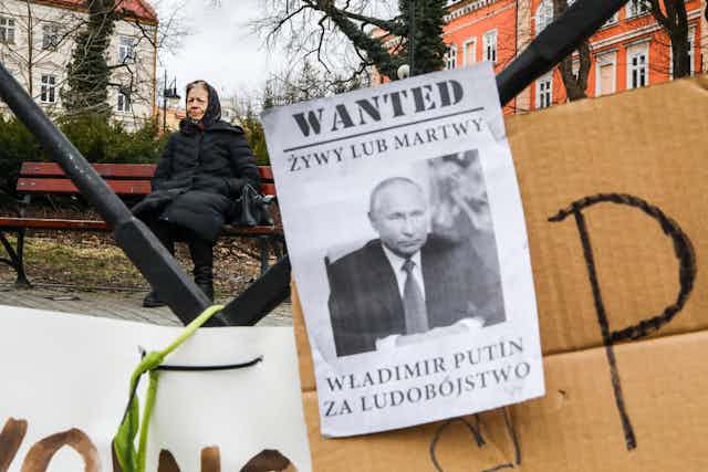 Banner reading 'Wanted Dead Or Alive Vladimir Putin For Genocide' is shown in front of an old woman sitting on a bench