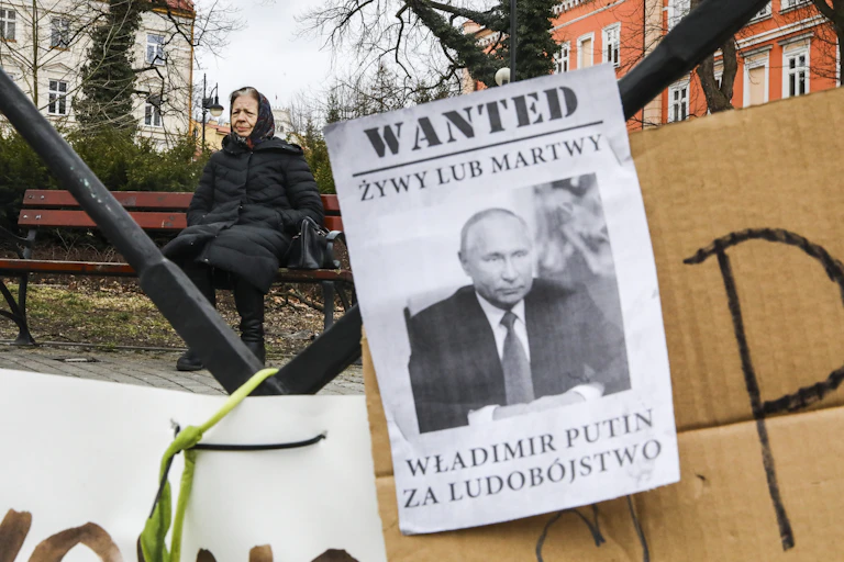 Banner reading 'Wanted Dead Or Alive Vladimir Putin For Genocide' is shown in front of an old woman sitting on a bench