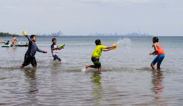 Children wade in a lake, throwing tubs of water at each other, with a city skyline in the background