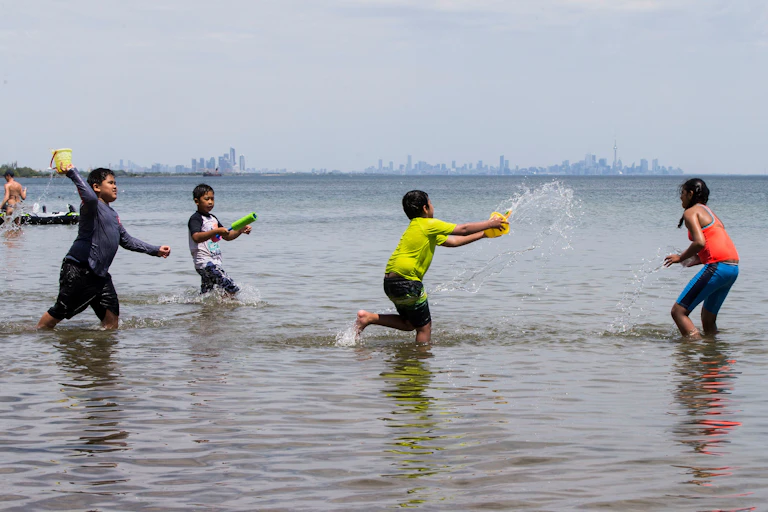 Children wade in a lake, throwing tubs of water at each other, with a city skyline in the background
