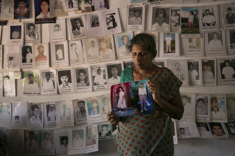 A woman holds two photographs in front of a wall of posters about missing people.