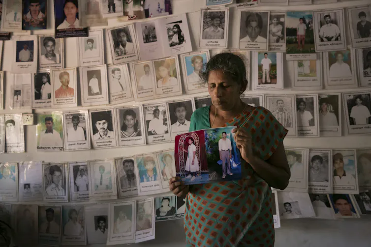 A woman holds two photographs in front of a wall of posters about missing people.