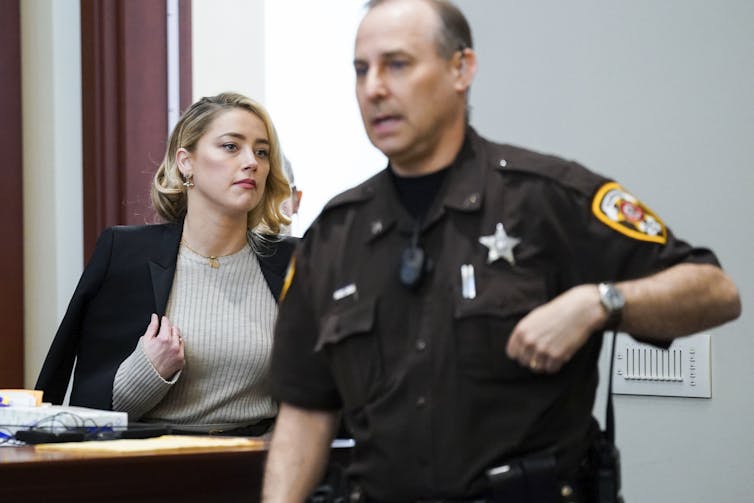 A woman with blond hear wears a sweater with a blazer over top, a man in sheriff uniform walks in front of her.