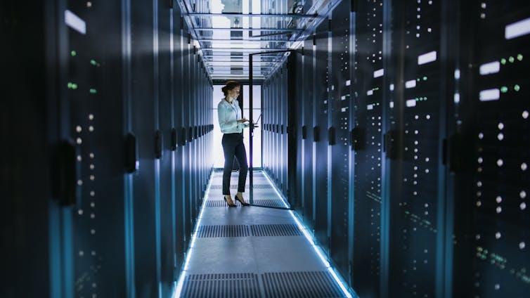 Server technician stands beside cabinet in data center hallway with rows of rack servers.  She is running diagnostics on her computer