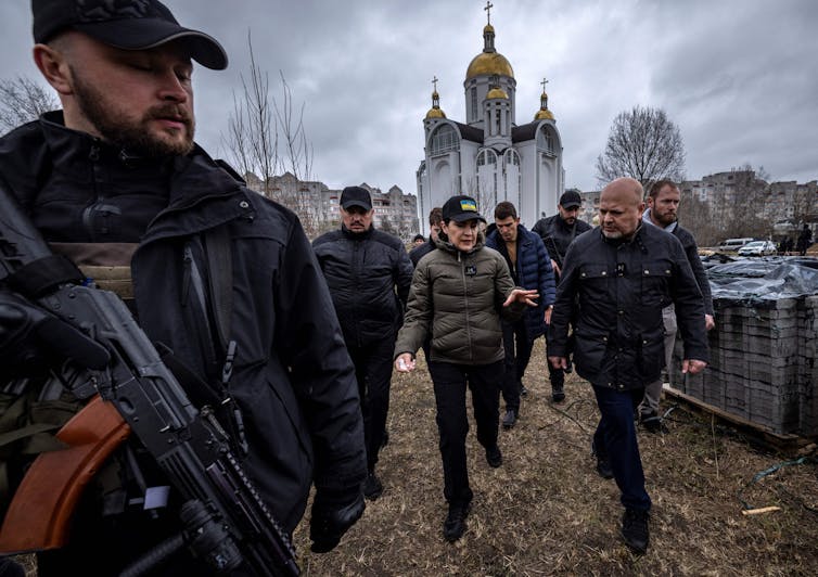 An armed guard stands in front of several people wearing dark clothes walking on a grey day. Behind them is a church.