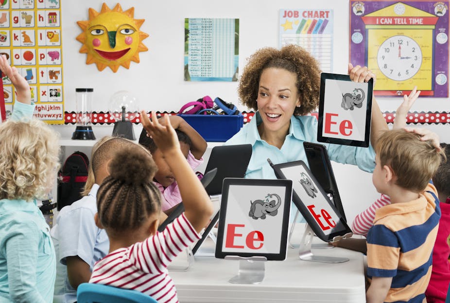 Several young children with iPads sit at a table as their teacher holds up an iPad with the letter "E" and a drawing of an elephant.