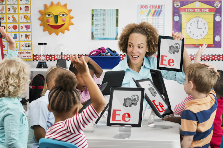 Several young children with iPads sit at a table as their teacher holds up an iPad with the letter "E" and a drawing of an elephant.