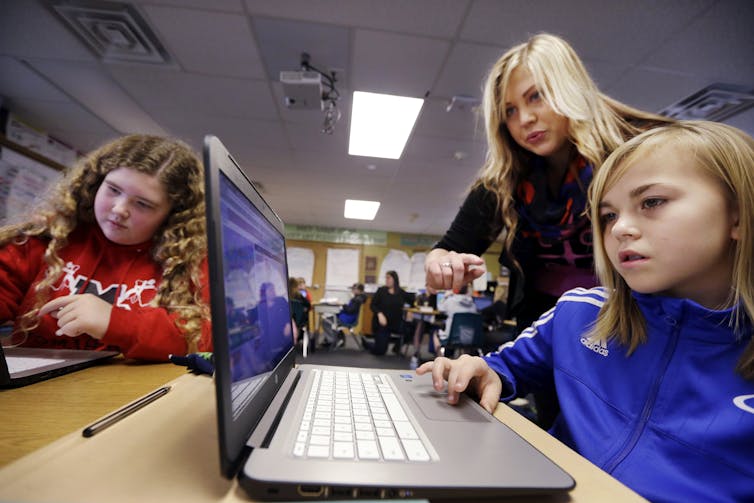 Gadis-gadis bekerja di komputer sementara seorang wanita membantu.