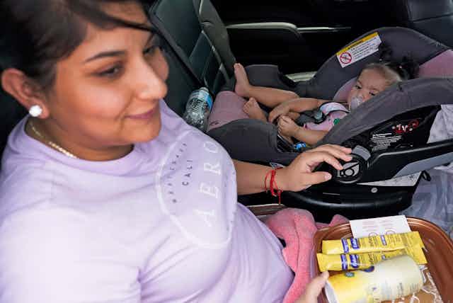 A woman sits in a car with baby formula on her lap next to her baby girl, sitting in a car seat with a pacifying in her mouth