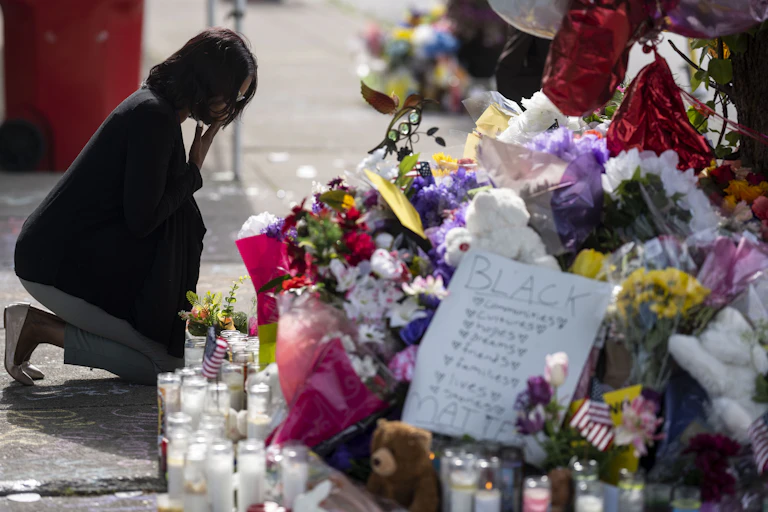 A woman with dark hair and dark jacket kneels in front of a large display of flowers and candles.