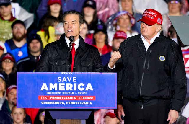 Donald Trump wearing a red 'Make America Great Again' hat looks on as Senate candidate Mehmet Oz speaks at a platform.