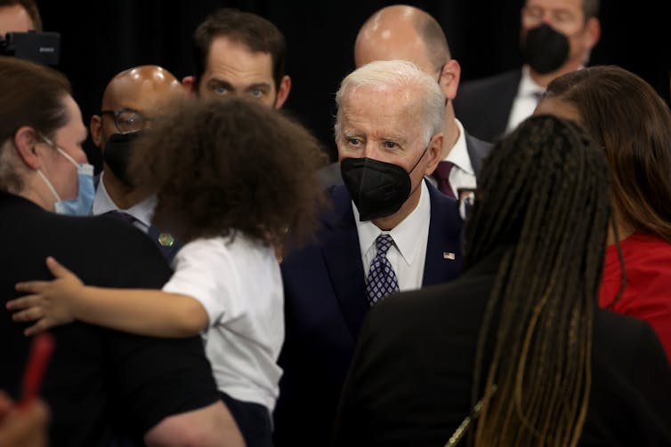 A white haired man in a black jacket and black face mask in a crowd of people.