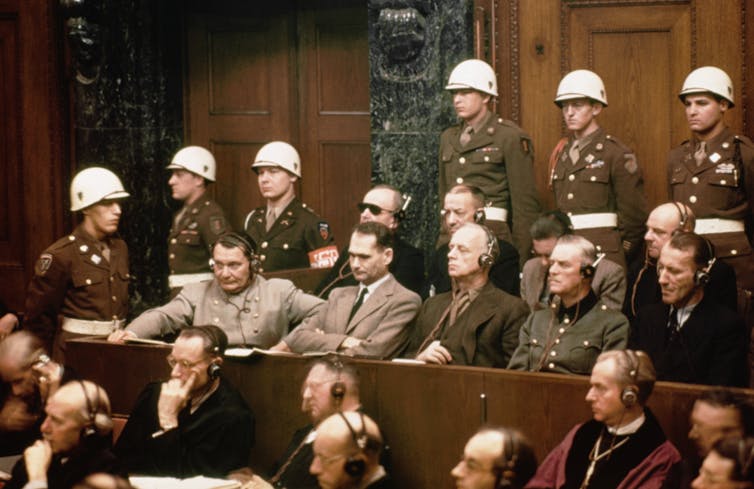 Dos filas de hombres están sentados en una sala de audiencias, rodeados por soldados con cascos blancos.