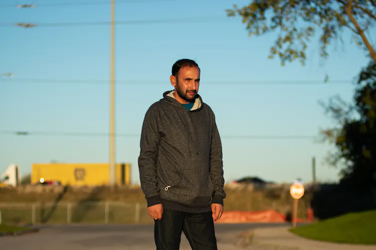 A dark-haired man with a beard stands on a quiet street.