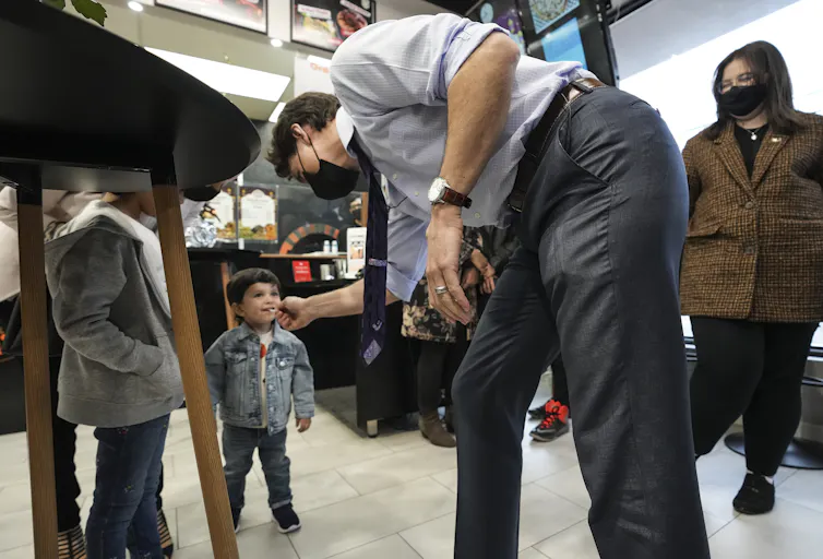 A man wearing a mask playfully reaches for the lollipop of a dark-haired toddler, who's smiling.