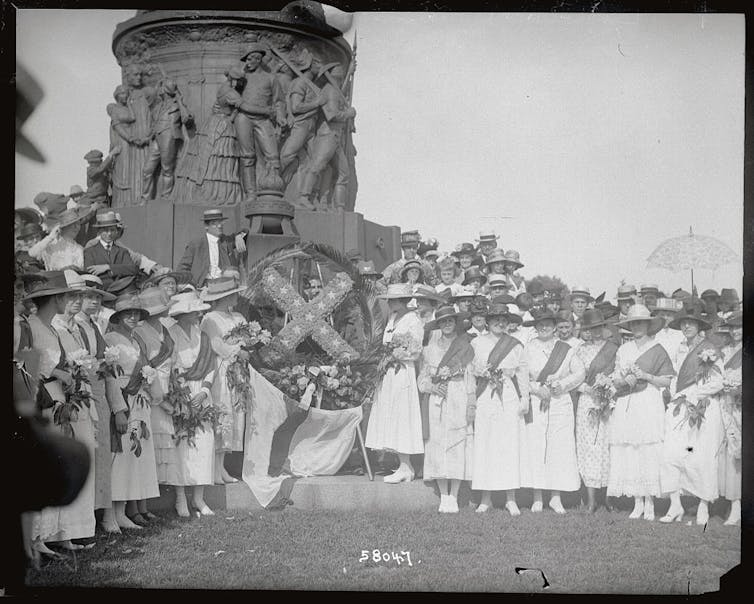 Women in white dresses and skirts stand in front of a war monument in a black and white photograph.
