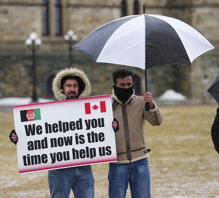 A man holds a sign that says 'We helped you and now is the time you help us.' Another man stands next to him under an umbrella.