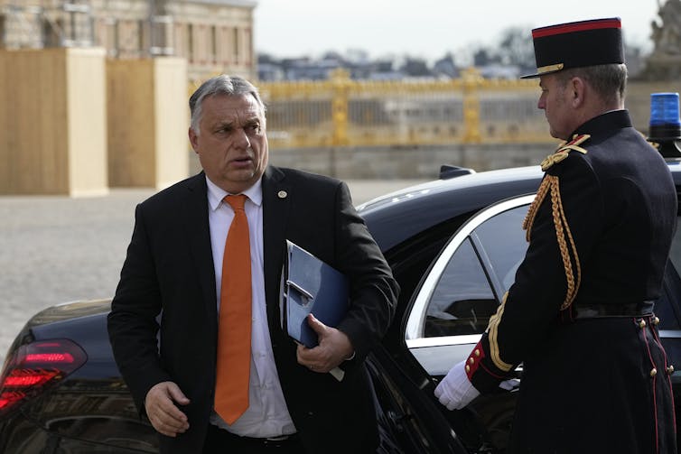 A man in a suit, carrying briefing books, steps out of a car as a uniformed guard holds the door open.