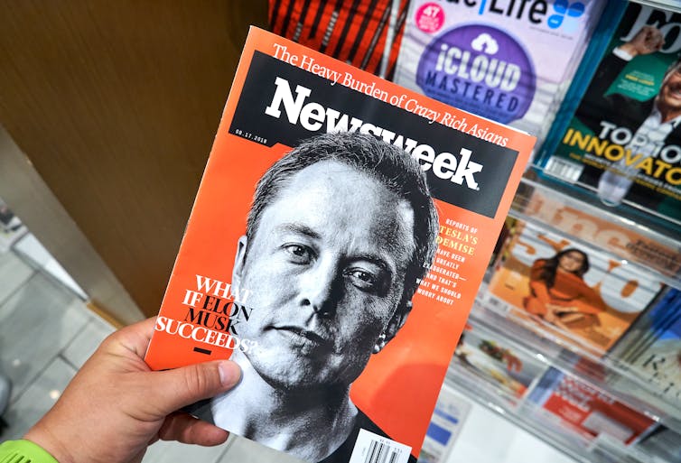 a hand holding a cover of Newsweek magazine, which has a black and white photograph of Elon Musk against an orange backdrop