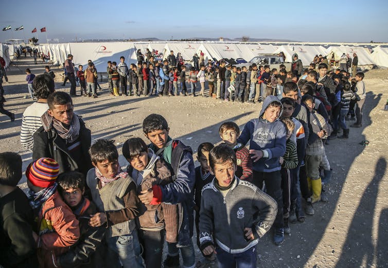 Syrian men and boys line up closely in a long row outside white tents.