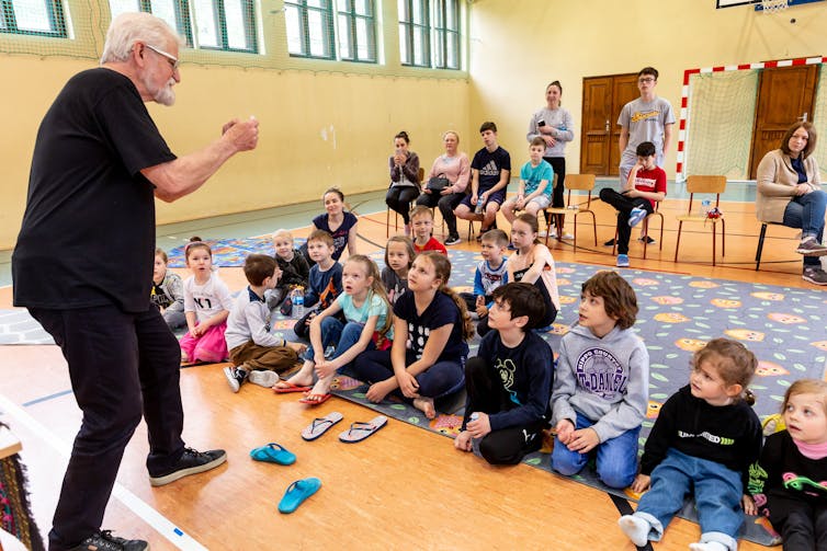 A man dressed in black with a white beard does a magic trick in front of a gym full of children, seated and watching.