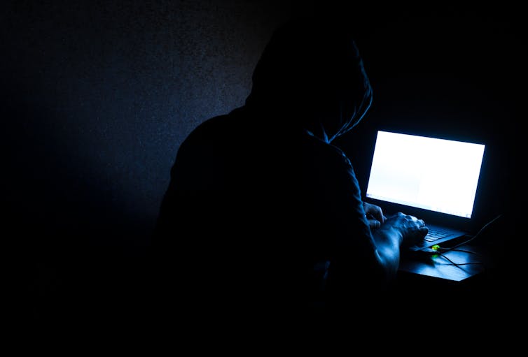 A darkened picture of a hooded man sitting alone at a laptop.