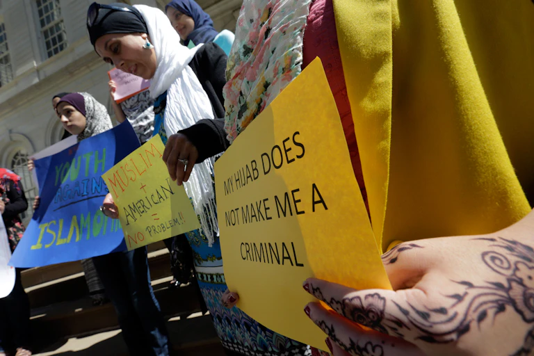 Women wearing head coverings, holding posters in protest, one of which says 'My hijab does not make me a criminal.'