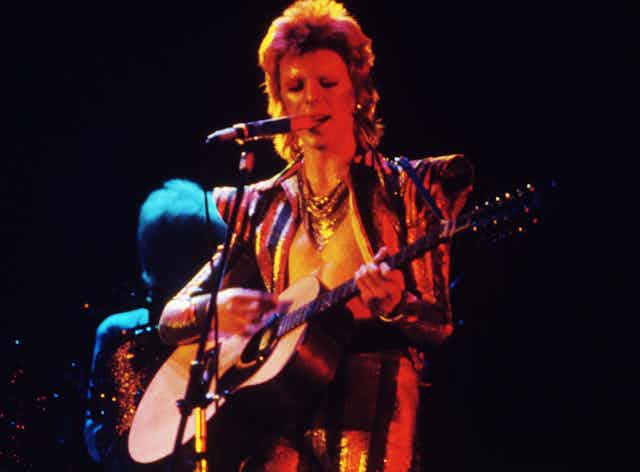 Man singing and playing guitar in 70s glam-rock attire.