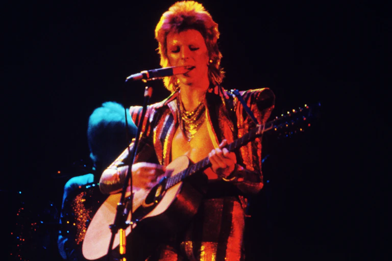 Man singing and playing guitar in 70s glam-rock attire.