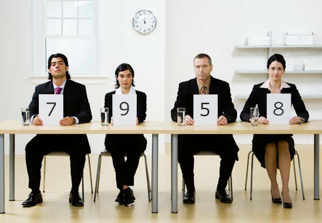 Four people wearing suits sit behind a table holding signs with different numbers on them