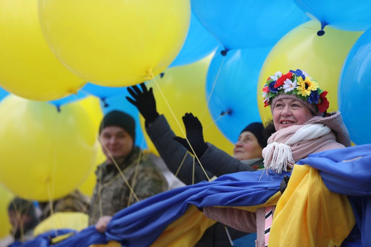 Women and men hold yellow and blue banner and yellow and blue balloons.
