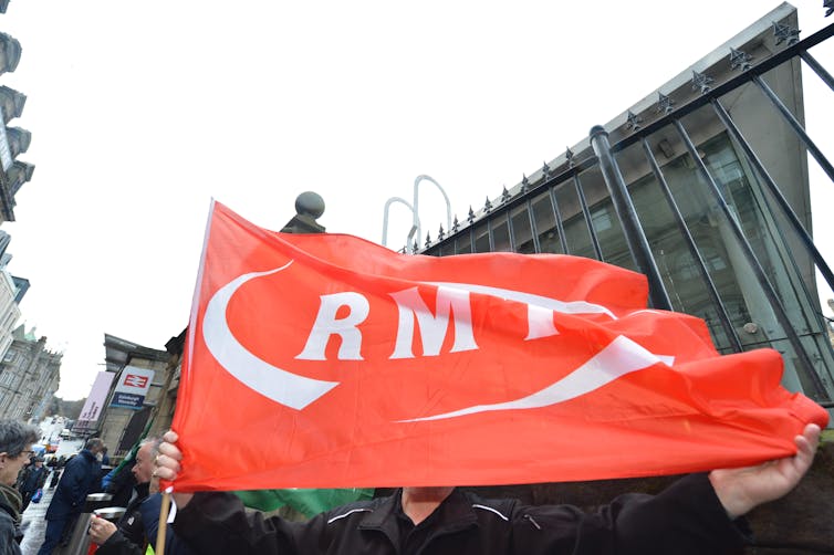 Striking rail workers hold up a large red flag in a street setting.