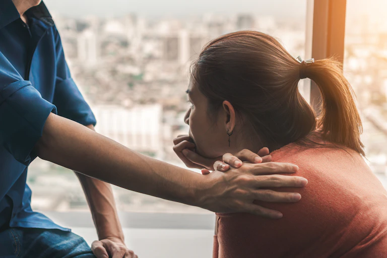 A woman facing away from the camera rests her chin on her hands while another person rests their hand on her shoulder
