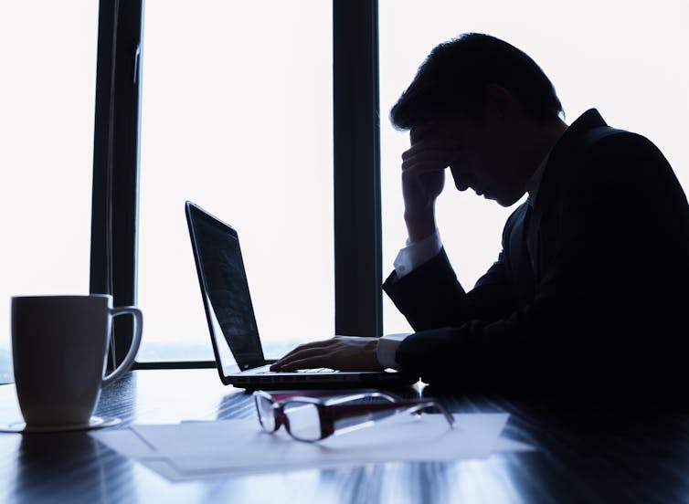Side view of a silhouette of a businessman at a laptop with his head resting on his hand and his glasses on the table next to him