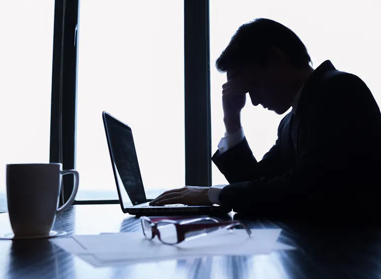 Side view of a silhouette of a businessman at a laptop with his head resting on his hand and his glasses on the table next to him