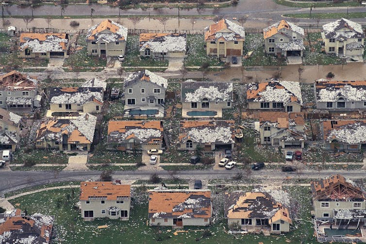 A neighborhood of homes with shredded roofs, some missing most of their roof tiles or shingles, others with parts of the roof missing entirely.